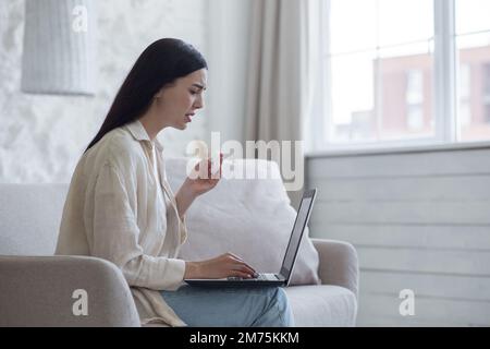 Aufgebrachte junge Frau sitzt zu Hause auf der Couch am Fenster mit einem Laptop, liest schlechte Nachrichten, erhielt eine E-Mail. Weinen, Tränen mit einer Serviette abwischen. Stockfoto