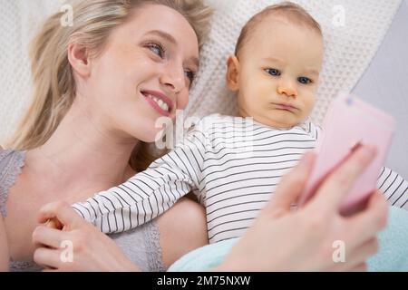 Eine Frau mit einem Baby, die ein Selfie macht und auf der Decke liegt Stockfoto