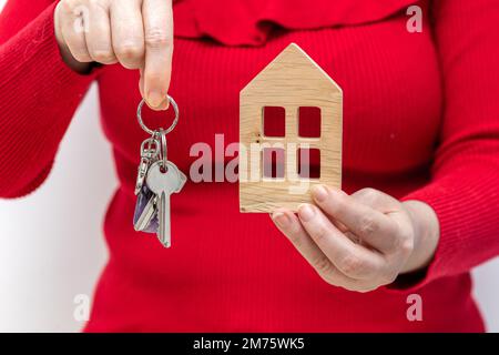 Wohnungskonzept, Eine Frau hält die Schlüssel ihres Traumhauses und eine hölzerne Miniatur einer Hütte in ihren Händen, Immobilienkauf Stockfoto