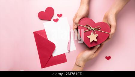 St. Valentinstagsbrief und Geschenke in herzförmiger Box in den Händen. Kopierbereich für Liebesbotschaft. Weiße Karte mit rotem Papierumschlag, Modell. Flach verlegt Stockfoto