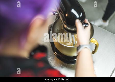 Konditorin POV. Eine Frau mit bunten Haaren, die einen Mixer benutzt und an der Theke in ihrer Schürze steht. Professionelle Bäckerei Küche. Hochwertiges Foto Stockfoto