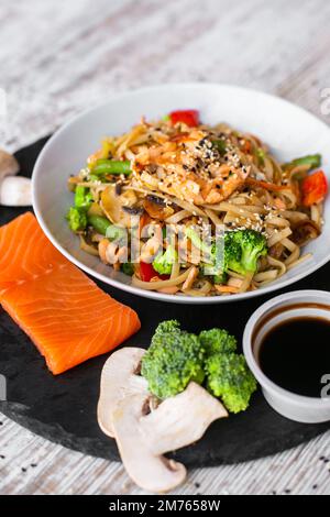 Wok, Nudeln, udon mit Lachs, auf schwarzem Teller und mit einem hölzernen weißen Hintergrund von oben Stockfoto