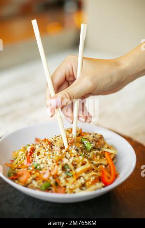 Chinesische Nudeln mit Garnelen und Gemüse. Eine Person, die Stäbchen mit Nudeln über frittiertem asiatischem udon hält. Stockfoto