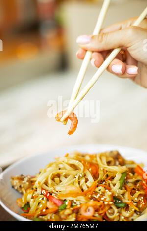 Chinesische Nudeln mit Garnelen und Gemüse. Eine Person, die Stäbchen mit Shrimps über asiatisch gebratenem udon hält. Stockfoto