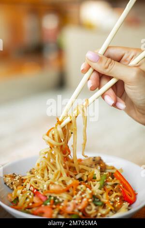 Chinesische Nudeln mit Garnelen und Gemüse. Eine Person, die Stäbchen mit Nudeln über frittiertem asiatischem udon hält. Stockfoto