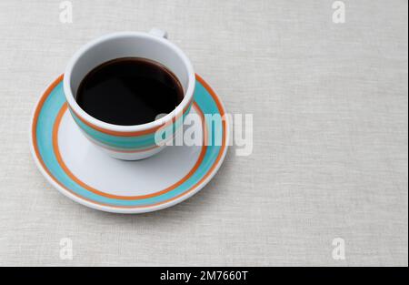 Eine bunte Tasse schwarzen Kaffees wird auf die Untertasse gestellt. Sowohl die Tasse als auch die Untertasse sind weiß mit orangefarbenen und aquafarbenen Ringen. Stockfoto