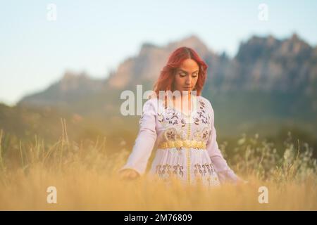 Eine wunderschöne rothaarige Dame mit einem gestickten weißen Kleid und einem goldenen Gürtel auf einem sonnigen Feld in Spanien Stockfoto