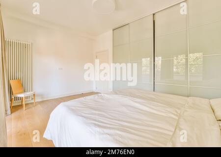 Ein Schlafzimmer mit einem Bett und Schiebetüren an der Wand vor dem Bett ist ein orangefarbener Stuhl neben dem Fenster Stockfoto