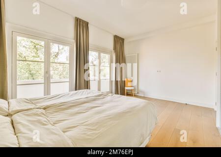 Ein Schlafzimmer mit weißen Wänden und Holzfußboden das Zimmer ist gut beleuchtet durch große Fenster mit Blick auf die Straße Stockfoto