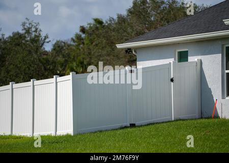 Weißer Lattenzaun aus Vinyl auf grünem Rasen, der das Grundstück umgibt, um den Garten zu schützen und Privatsphäre zu genießen Stockfoto