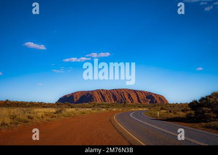 Outback, Australien - 12. November 2022: Straße zum berühmten Wahrzeichen des australischen Outbacks. Der rote Felsen des Uluru oder Ayres. Blauer Himmel Wüste la Stockfoto