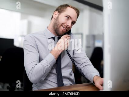 Er ist ein engagierter junger Profi. Ein gutaussehender junger Mann, der den Computer benutzt, während er bei der Arbeit telefoniert. Stockfoto