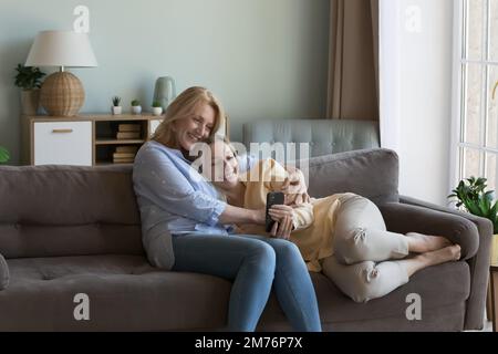 Fröhliche, reife mutter, die junge Erwachsene, hübsche Tochter mit Smartphone kuschelt Stockfoto