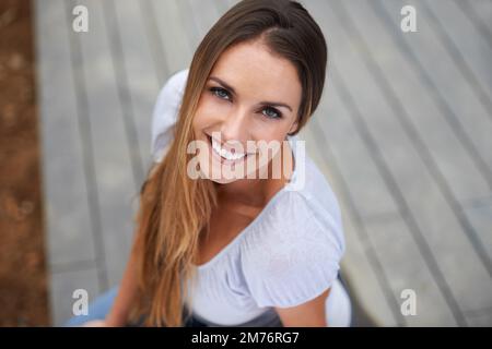 Vergessen Sie nicht zu lächeln. Ein Hochwinkel-Porträt einer schönen Frau, die vor der Kamera lächelt, während sie draußen sitzt. Stockfoto