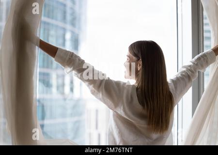 Die Frau öffnet die Vorhänge, begrüßt den neuen Tag, genießt den Blick auf die große Stadt Stockfoto