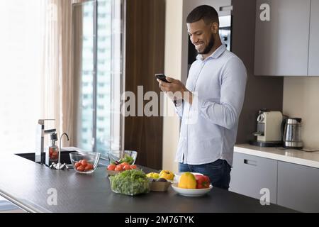 Junger, gutaussehender Afrikaner, der Handy benutzt und in der modernen Küche steht Stockfoto