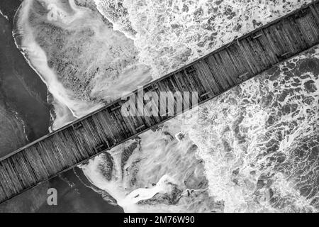 Ein Blick aus der Vogelperspektive auf einen hölzernen Pier auf Meereswellen, die in Graustufen gegen den Strand schlagen Stockfoto
