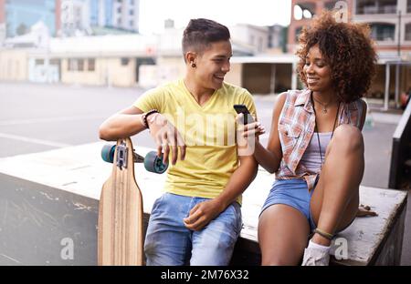Zusammen in der Stadt abhängen. Ein junges Paar, das sich auf dem Handy etwas ansieht, während es Skateboard fährt. Stockfoto