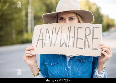 Portrait Nahaufnahme einer Frau, die hoffentlich am Auto vorbeifährt, mit einem Pappposter am Straßenrand. Frau mit Hut Flucht aus der Stadt mit Auto-Stopp nach Stockfoto