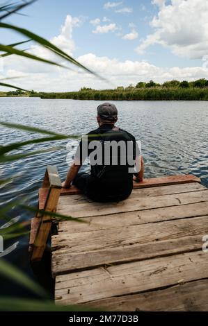 Ein Mann sitzt am Rand einer hölzernen Brücke in der Nähe eines Sees neben Schilf, einem tiefen und klaren Gewässer am Horizont. Stockfoto