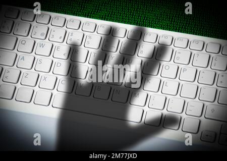 Ein bedrohlicher Handschatten auf einer Computertastatur vor gedruckten Computerdaten. Stockfoto