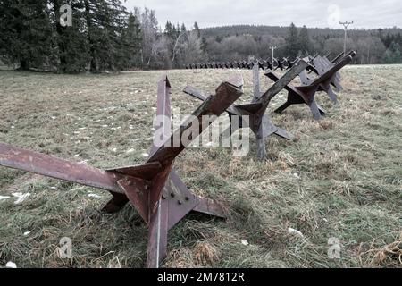 An einem düsteren, dunklen Wintertag ohne Schnee lagen alte, rostige Panzerabwehrsperren aus Stahl auf Gras mit Wald im Hintergrund. Konzept für Krieg, Verteidigung Stockfoto