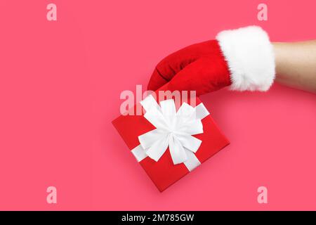 Der Weihnachtsmann in roten Handschuhen hat ein Geschenk in der Hand. Rote Box mit einer Schleife in der Hand des Weihnachtsmanns, isoliert auf rosa Hintergrund. Weihnachten und Neujahr Stockfoto