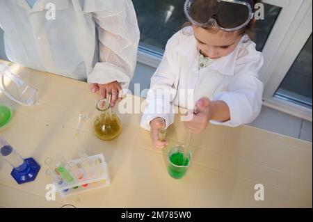 Draufsicht eines intelligenten Schulmädchens mit einer graduierten Pipette, die wenige Reagenzien in einen Becher mit chemischer Lösung tropft Stockfoto