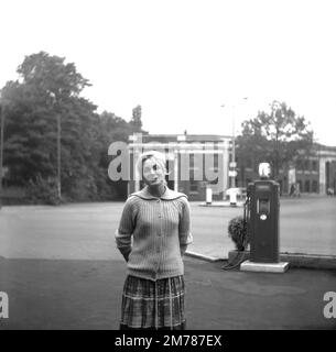 1950er, historisch, junge Frau, die für ihr Foto auf dem Vorplatz von Hargood Motor Co (Parrs Wood) Ltd, Didsbury, England, Großbritannien steht. Hinter ihr eine Zapfsäule der Ära, vielleicht ein Regente. Der Busbahnhof, der den Großraum Manchester anfährt, ist im Hintergrund zu sehen Stockfoto