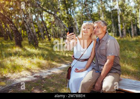 Porträt eines älteren Familienpaares, das Selfie mit dem Smartphone im Sommerwald macht. Ältere Menschen ruhen sich auf einer Bank aus Stockfoto