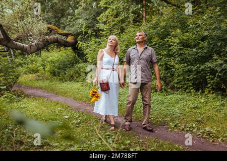 Stilvolles älteres Familienpaar, das im Sommerwald draußen spaziert. Ältere Menschen, die Händchen halten. Die Frau trägt Blumen Stockfoto
