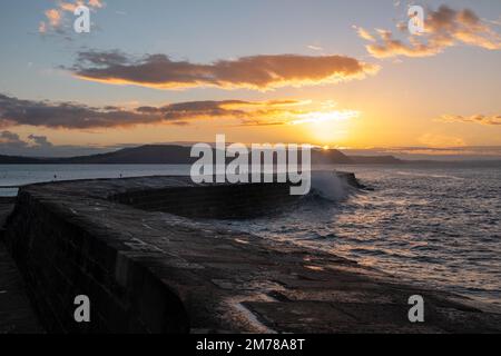 Die Welle stürzt über das Ende des Cobb Dorset England mit einem strahlend orangefarbenen Sonnenaufgang über dem Meer im Hintergrund Stockfoto