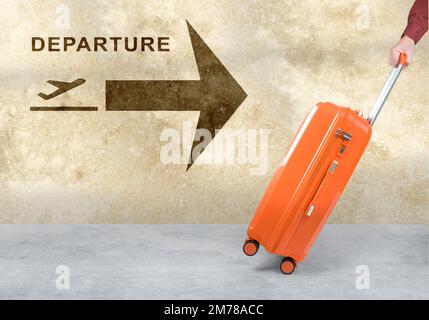 Hand hält den orangefarbenen Koffer auf weißem Hintergrund. Flughafenschild mit Abflug Flugzeugsymbol an der Wand. Rollendes Gepäck des Passagiers in Bewegung. Sommerurlaub Stockfoto