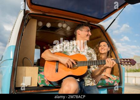 Foto eines Paares aus einem niedrigen Blickwinkel, das Gitarre spielt, auf einem Roadtrip mit einem Retro-Van Stockfoto