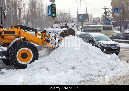 Die Klinge eines kommunalen Traktors entfernt an einem Wintertag während eines Schneefalls Schneefälle von einer Straße zwischen den Straßen der Stadt. Stockfoto