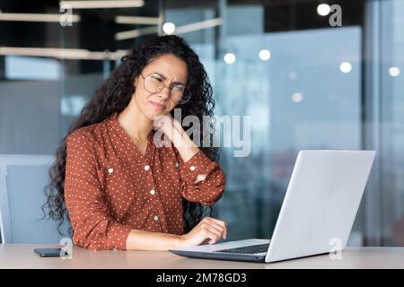 Müde, junge lateinamerikanische Geschäftsfrau, Freiberufler. Er sitzt am Schreibtisch im Büro, arbeitet Überstunden am Laptop. Hält und massiert den Hals mit der Hand, fühlt Schmerzen, Spannung. Stockfoto