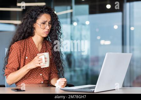 Eine kranke junge Geschäftsfrau aus Lateinamerika, eine Freiberuflerin, sitzt am Schreibtisch im Büro und arbeitet an einem Laptop. Er hält eine Tasse Tee und eine Serviette in den Händen. Sie ist aufgebracht, sie fühlt sich schlecht. Stockfoto