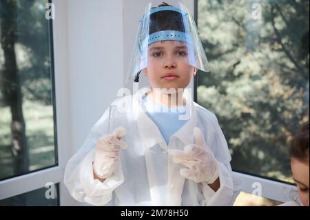 Ein gutaussehender, kluger Teenager, der Laborkleidung trägt, Reagenzgläser hält, selbstbewusst in die Kamera schaut Stockfoto