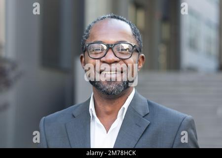 Nahaufnahme. Portrait eines erfolgreichen, gutaussehenden afroamerikanischen Mannes. Ein Geschäftsmann, Anwalt, Boss steht im Anzug und in der Brille vor dem Büro, schaut in die Kamera und lächelt. Stockfoto