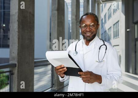 Porträt eines gutaussehenden afroamerikanischen Arztes. Er steht neben dem Krankenhaus in einem weißen Mantel mit einem Ordner in den Händen, schaut in die Kamera und lächelt. Stockfoto