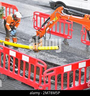 Die Hydraulikschelle des Mini-Grabers ist am hinteren Trägerarm befestigt und greift 125mm neue gelbe Gasleitung aus Kunststoff, die über der Grube gehalten wird und an einem Punkt in England zugeschnitten wird Stockfoto