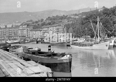 Schlepper und Frachtschiffe oder Holzschiffe mit Holzstämmen im Hafen oder Hafen mit der Anlegestelle und dem Hafengebiet Bergen Norwegen c1960. Klassische Schwarzweiß- oder Schwarzweißfotos. Stockfoto