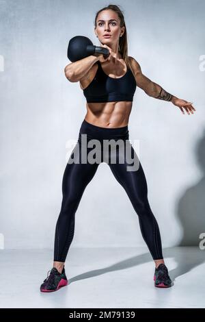 Sportliches Frauen-Workout mit Kugelhantel. Foto einer Frau in schwarzer Sportbekleidung auf grauem Hintergrund. Sportliche Motivation und gesunder Lebensstil Stockfoto