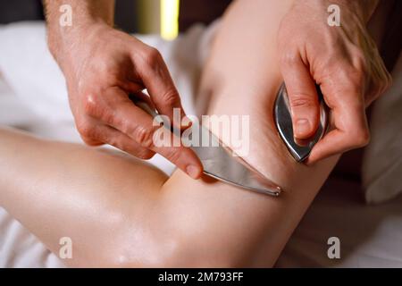 Nahaufnahme der Hände eines Masseurs, der eine iastme Massage mit einem speziellen Edelstahlwerkzeug für die Frau im Spa-Salon durchführt Stockfoto