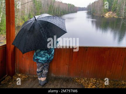 Eine Frau steht unter einem Schirm auf einer hölzernen Plattform am Ufer des Sees Stockfoto