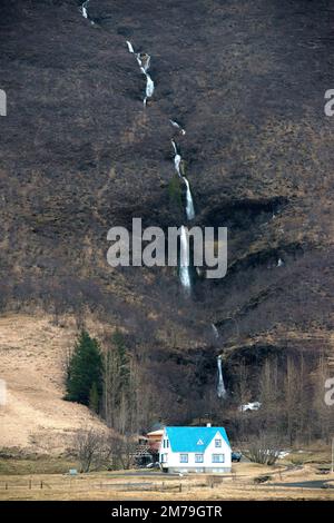 Ein kleiner Wasserfall stürzt einen Berg hinunter mit einem blauen Haus im Vordergrund, in island, abseits der Ringstraße, Route 1. Stockfoto