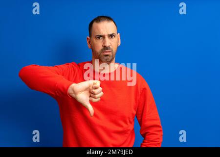 Bärtiger lateinamerikanischer Mann mit rotem Pullover schmollt mit dem Daumen nach unten, mit negativer Einstellung, isoliert auf blauem Hintergrund. Stockfoto