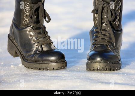 Weibliche Beine in schwarzen Leder Schnürstiefeln auf Schnee. Frau, die auf der Winterstraße läuft, warme Schuhe für kaltes Wetter Stockfoto