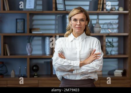 Porträt einer leitenden, seriösen Chefin im klassischen Büro am Abend, einer reifen Geschäftsfrau mit gekreuzten Armen, die aufmerksam vor die Kamera schaut Stockfoto