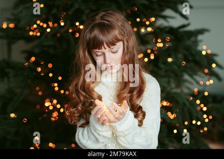 Glückliches junges Mädchen, das mit Girlande am Weihnachtsbaum sitzt Stockfoto